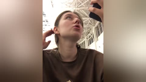 Media: A video of a young woman with light skin and brown hair in a dark brown t-shirt, standing indoors, looking upwards with a contemplative expression.