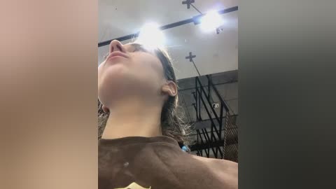 Media: Video of a young woman with light skin and dark hair, wearing a grey shirt, standing with her head tilted back in a gym with bright lights and exercise equipment in the background.