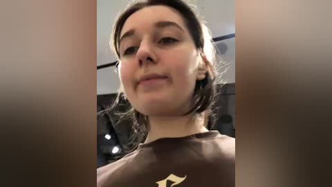 Media: A video of a young woman with fair skin and brown hair, wearing a brown shirt, taken from a low angle, showing her from the chest up. The background is blurry.