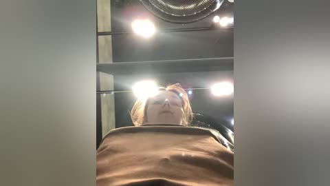 Media: Video of a person with short blonde hair, wearing a brown shirt, sitting in a car with bright headlights shining directly into the camera.