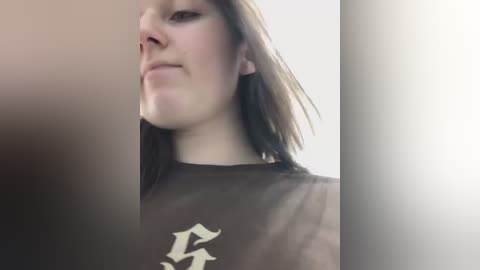 Media: Video of a young woman with light skin and shoulder-length brown hair, wearing a black shirt with a white logo. The background is blurred, with a soft focus on the subject.