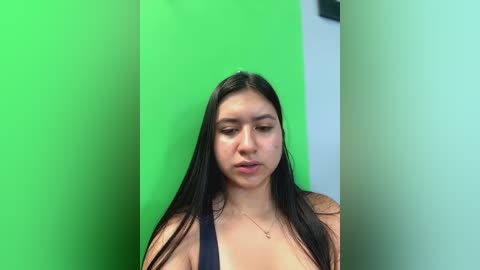 Media: Video of a young Latina woman with long, straight black hair, wearing a black top, standing in a bright green room. She has a neutral expression, and the image is slightly out of focus.