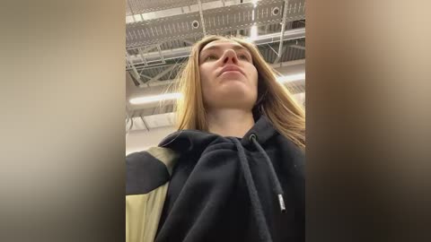 Media: Video of a young woman with long, straight, light brown hair, wearing a black hoodie, standing in a dimly lit room with exposed ceiling ducts.