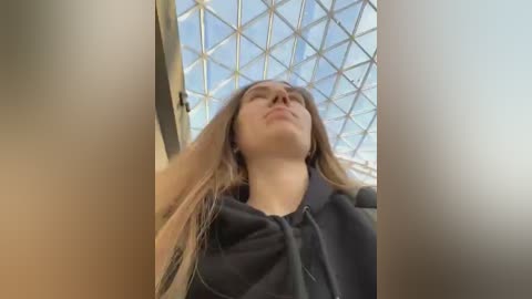 Media: Video of a young woman with long, straight blonde hair, wearing a black hoodie, standing in a modern glass atrium with a diamond-shaped glass ceiling. She has a confident, slightly smug expression.