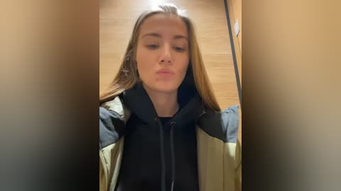 Media: Video of a young Caucasian woman with long, straight, light brown hair, wearing a black hoodie, and a beige jacket, making a kissy face while standing in a dimly lit hallway with light wooden floors.