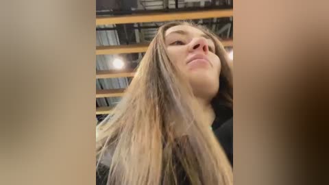 Media: Video of a young woman with long, straight, light brown hair, tilted head, and closed eyes. She is indoors, with blurred background showing industrial elements like metal beams and pipes.