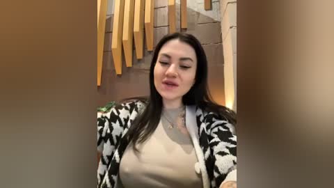 Media: Video of a smiling woman with long dark hair, wearing a patterned cardigan over a beige top, in a rustic setting with wooden shelves and bamboo hanging.