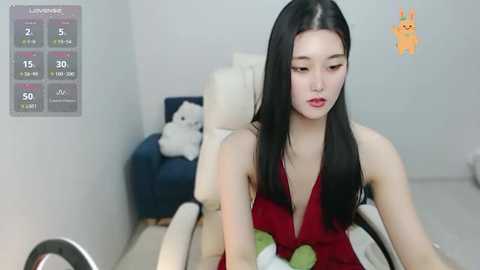 Media: Video of an East Asian woman with long black hair, fair skin, and red lipstick, wearing a sleeveless red dress, sitting on a beige couch with a white bear toy.