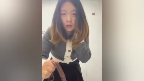 Media: Video of an Asian woman with long, straight, dark brown hair and fair skin, wearing a gray cardigan over a white blouse and black skirt, holding a brown bag. She has a startled expression.