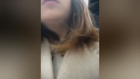 Media: A close-up video of a woman with fair skin, wearing a beige coat, her light brown hair partially covering her face, with blurred background.