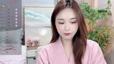 Media: Video of an East Asian woman with long, straight brown hair, wearing a pink bathrobe, standing indoors. Background includes a digital clock and a potted plant.