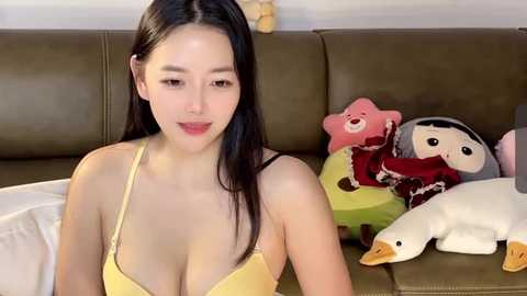 Media: Video of a young Asian woman with long black hair, wearing a yellow bra, sitting on a green couch with stuffed animals.