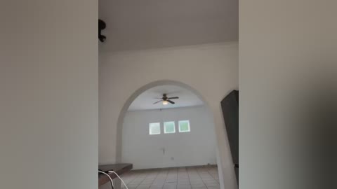 Media: Video of an unfinished, spacious room with white walls, an arched doorway leading to a kitchen, wooden floor, and a ceiling fan.