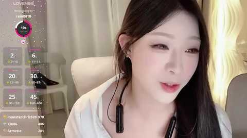 Media: Video of an East Asian woman with long black hair, wearing a white top, headphones, and makeup, in a cozy indoor setting with a light-colored curtain and a soft chair.