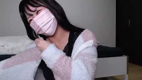 Media: Video of an Asian woman with long black hair, wearing a pink mask, black top, and a white and pink fuzzy robe, sitting on a bed with white sheets and black blankets.