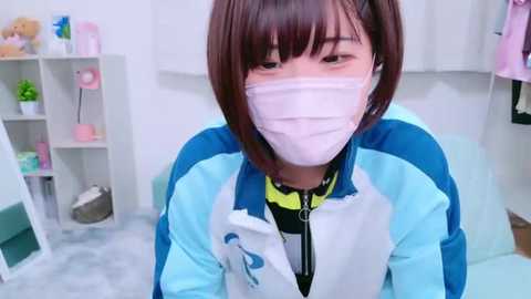 Media: A video of an Asian woman with short brown hair, wearing a white face mask, a blue and white jacket, and a black shirt, sitting in a white room with shelves filled with toys and plants.