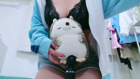 Media: Video of a woman in a black lace bra, blue jacket, and denim shorts, holding a plush hedgehog. Background includes hanging clothes.