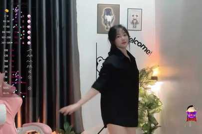 Media: Video of a slim, fair-skinned woman with black hair wearing a black robe, dancing in a modern room with black curtains, framed art, and a green plant.