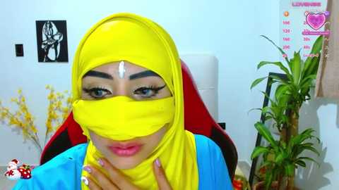 Media: A video of a woman in a yellow hijab and face mask, sitting in a red chair, surrounded by a white room with a plant and a poster.