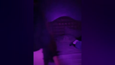 Media: Video with a purple tint, featuring a blurred figure in a dimly lit room. The figure appears to be a person with dark hair, partially obscured by shadows.