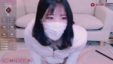 Media: A video of an Asian woman with long black hair, wearing a white mask, kneeling in a living room with a beige couch and a pink rug.