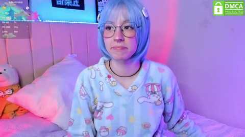 Media: Video of a young woman with blue hair, wearing glasses, and a cartoon pajama set, sitting on a bed with plush toys and a colorful background.