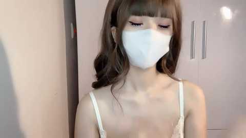 Media: Video of a young woman with fair skin, wearing a white face mask, white lace bra, and long brown hair. She stands in front of a white wardrobe, with a blurred background.