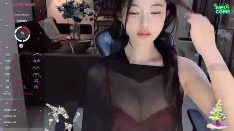 Media: A video of a young Asian woman with fair skin, wearing a sheer black top that reveals her bra, sitting in a modern living room with a gaming setup, playing a video game.