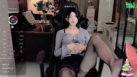 Media: Video of a woman in a gaming chair, wearing a grey crop top, black shorts, and thigh-high stockings, with a relaxed expression, in a modern living room.