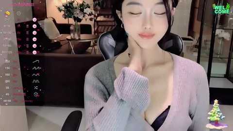 Media: A video of a young Asian woman with fair skin, wearing a low-cut grey sweater, sitting in a modern living room with a TV and sofa in the background.