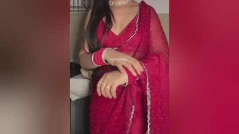 Media: Video of a woman in a red sari, adorned with intricate white embroidery, standing against a muted beige backdrop. Her long, dark hair cascades over her shoulders.