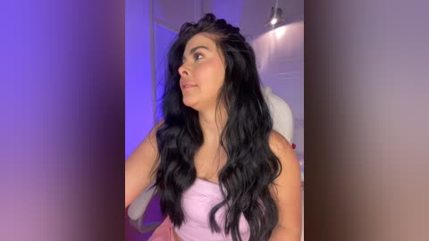 Media: Video of a young woman with long, wavy black hair, wearing a light pink top, standing in a softly lit room with purple and blue lighting accents.