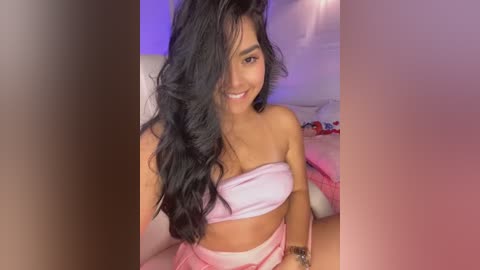 Media: Video of a smiling Latina woman with long, wavy black hair, wearing a pink strapless top and high-waisted shorts, sitting on a bed with pink sheets and pillows, bathed in soft purple lighting.