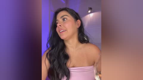 Media: Video of a smiling, light-skinned woman with long, wavy black hair, wearing a strapless pink top. Background features a dimly lit room with purple lighting.