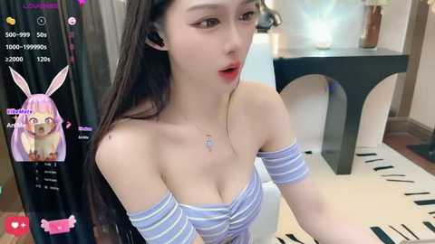 Media: A video of a young Asian woman with long, straight black hair and light skin, wearing a striped off-shoulder top, sitting on a black armchair in a modern room with a piano.