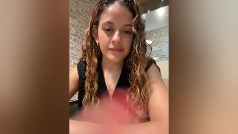 Media: A video of a young woman with light skin, curly brown hair, and a black sleeveless top, smiling, indoors with brick walls and modern lighting.