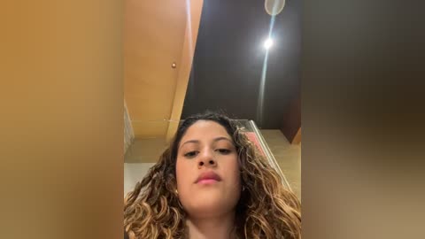 Media: A video of a young woman with medium brown skin and curly brown hair, wearing minimal makeup, standing in a dimly lit room with beige walls, a wooden door, and a ceiling light.