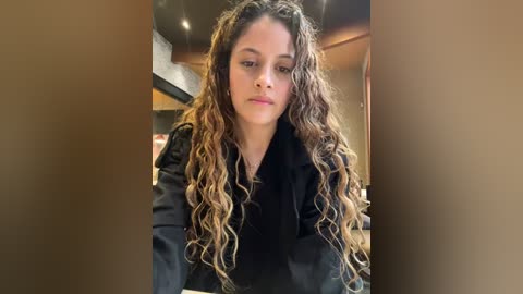 Media: Video of a young woman with long, curly, wavy brown hair and fair skin, wearing a black jacket, indoors with dim lighting and blurred background.