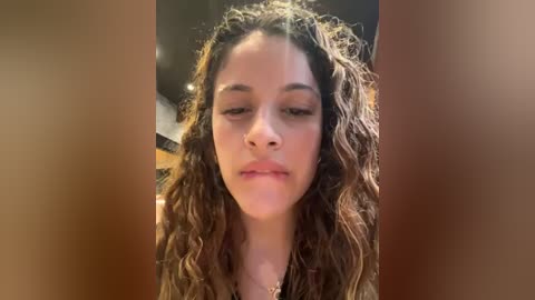 Media: Video of a young woman with curly brown hair, light skin, and closed eyes, wearing a gold necklace, in an indoor setting with blurred background.