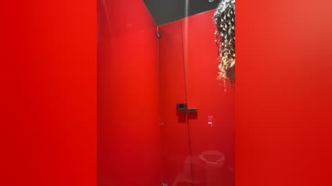 Media: A video of a small, modern restroom with vivid red walls and a glass shower enclosure. The showerhead is attached to the wall, and a curly, blonde wig hangs from a hook above the toilet.