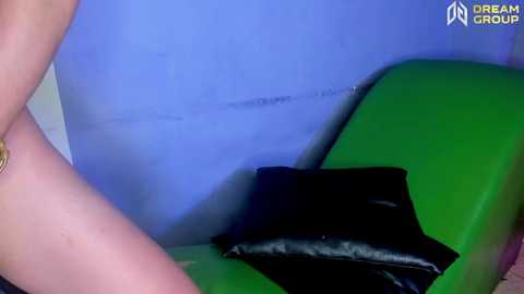 Media: Video of a woman with fair skin and a gold bracelet, sitting on a green couch with a black pillow, against a blue wall.