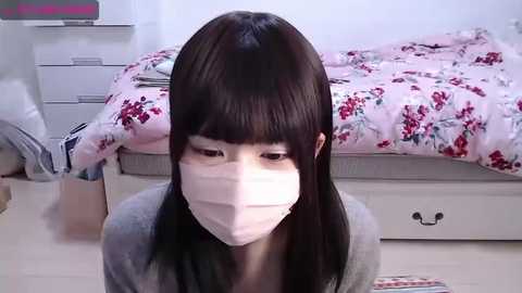 Media: Video of an Asian woman with straight black hair and a white face mask kneeling in a tidy bedroom with floral bedding, white drawers, and a small stool.