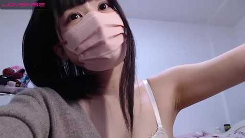 Media: A video of an Asian woman with long black hair and a pink face mask, wearing a white bra and gray cardigan, in a dimly lit room with cluttered background.