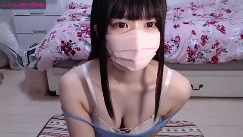 Media: Video of an Asian woman with long black hair, wearing a pink mask and a blue lace bra, kneeling on a striped rug in a pink floral bedroom.