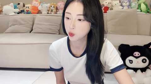 Media: Video of an East Asian woman with long black hair, wearing a white T-shirt, blowing a kiss, surrounded by stuffed animals in a cozy living room.