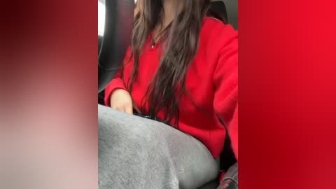 Media: Video of a woman with long brown hair, wearing a red sweater, seated in a car, with a blurred red background.