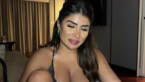 Media: Video of a curvy, light-skinned woman with long dark hair, wearing a black halter top, smiling and looking down. Background features beige curtains, a flat-screen TV, and a wooden table.