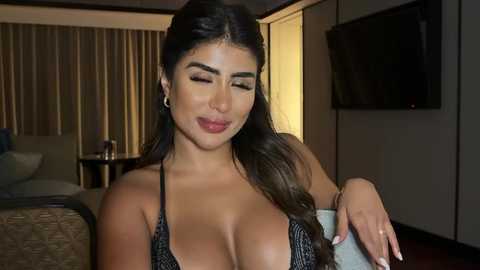 Media: Video of a curvy, light-skinned Latina woman with long, dark hair, wearing a black lace bralette, smiling in a modern, dimly-lit bedroom with a flat-screen TV on the wall.