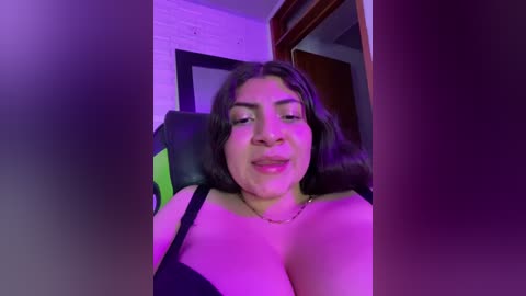 Media: Video of a Latina woman with medium skin tone, long dark hair, wearing a black bra, seated on a black gaming chair, illuminated by purple lighting, indoors.