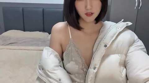 Media: Video of an East Asian woman with straight black hair, fair skin, wearing a white puffer jacket over a lacy grey bra, sitting on a bed with beige sheets.